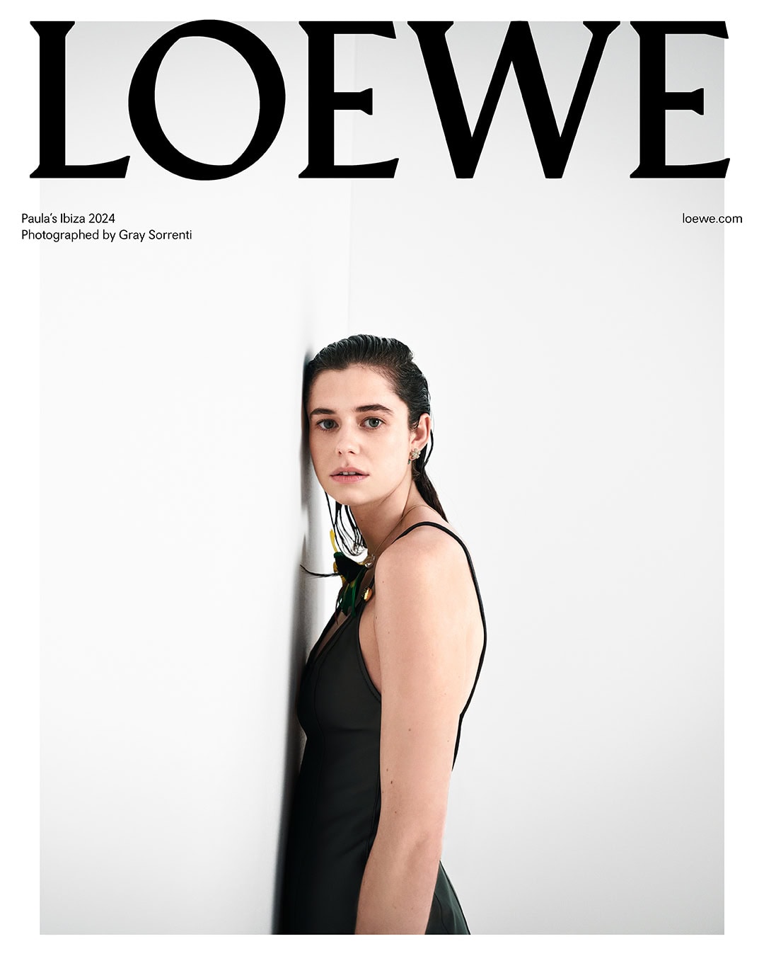 Fashion editorial shot of a female model in a dark dress leaning against a white wall, promoting Loewe's Paula's Ibiza 2024 collection.