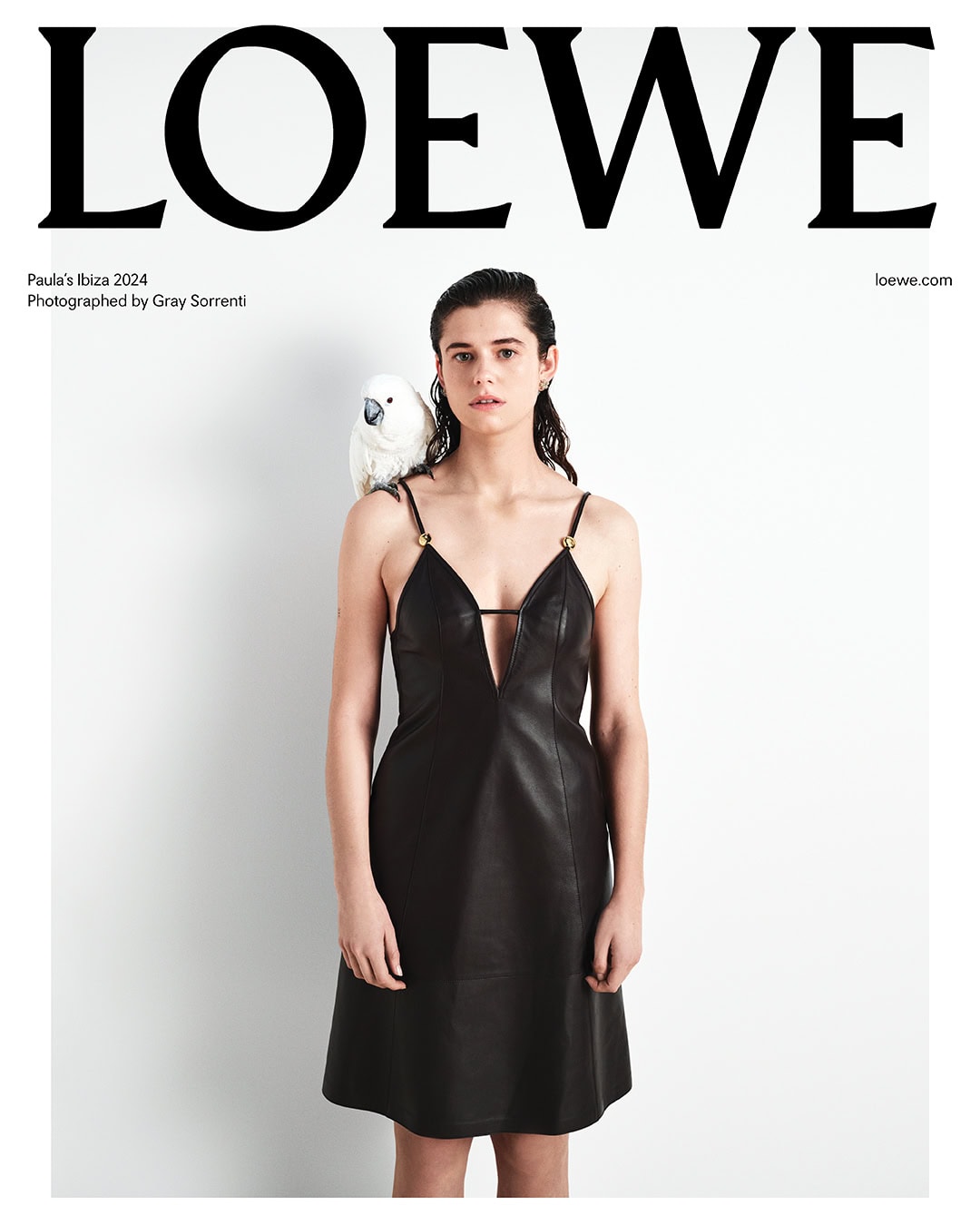 Fashion editorial featuring a woman in a black dress with a white cockatoo perched on her shoulder against a white background.