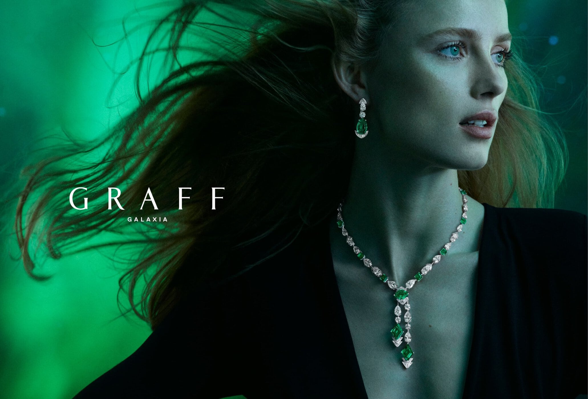 Elegant adult female wearing luxurious jewelry (necklace and earrings) posing against a blurred green bokeh background in an ad.