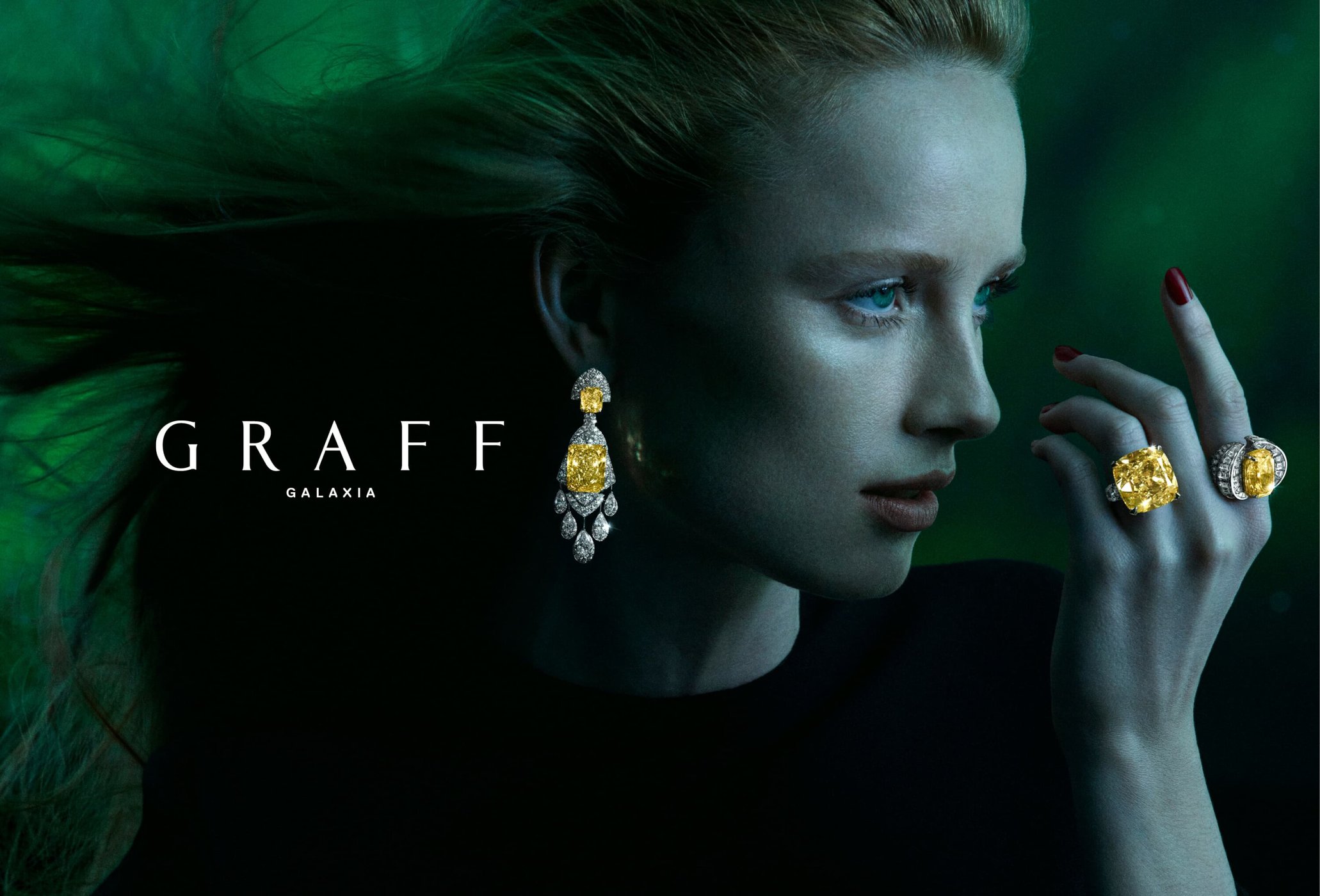 Blonde woman with yellow diamond earrings and rings in a jewellery advertisement, highlighted against a dark green background.