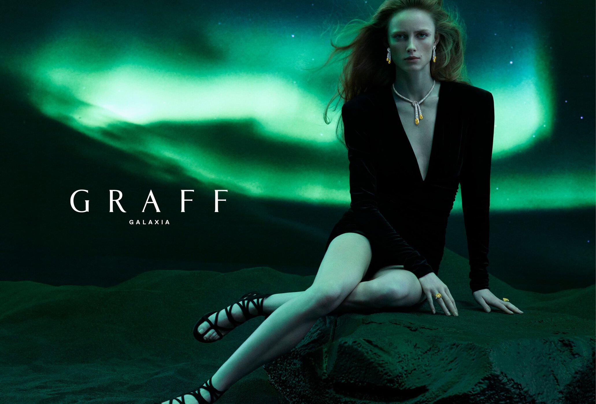 A fair-skinned woman wearing a black dress and Graff jewelry poses against an aurora borealis-style backdrop in a studio shot.