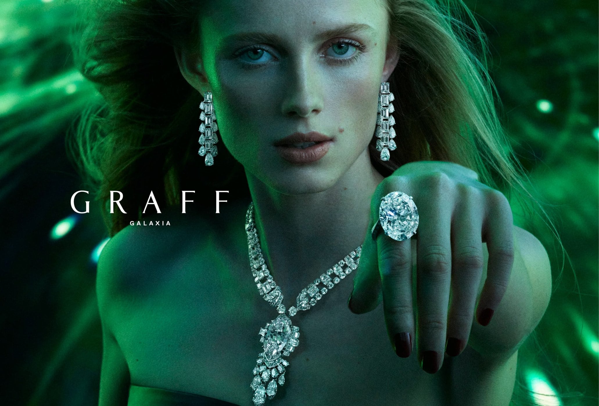 A woman is showcasing sparkling Graff jewelry, including a ring, earrings, and necklace, against a green background in a close-up shot.