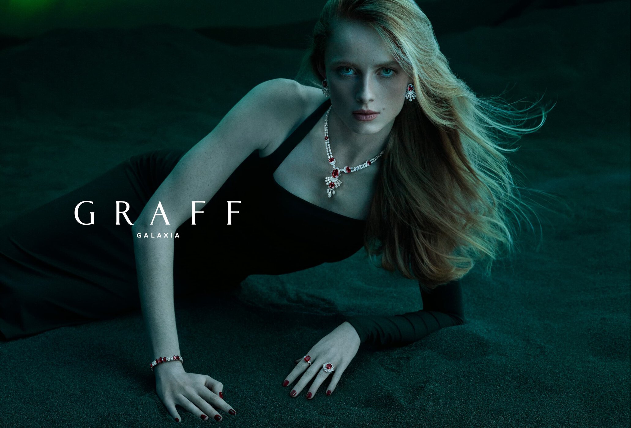 Elegant red-haired model in a black dress lying on dark sand, wearing a diamond necklace, earrings, ring, and bracelet for Graff jewelry ad.