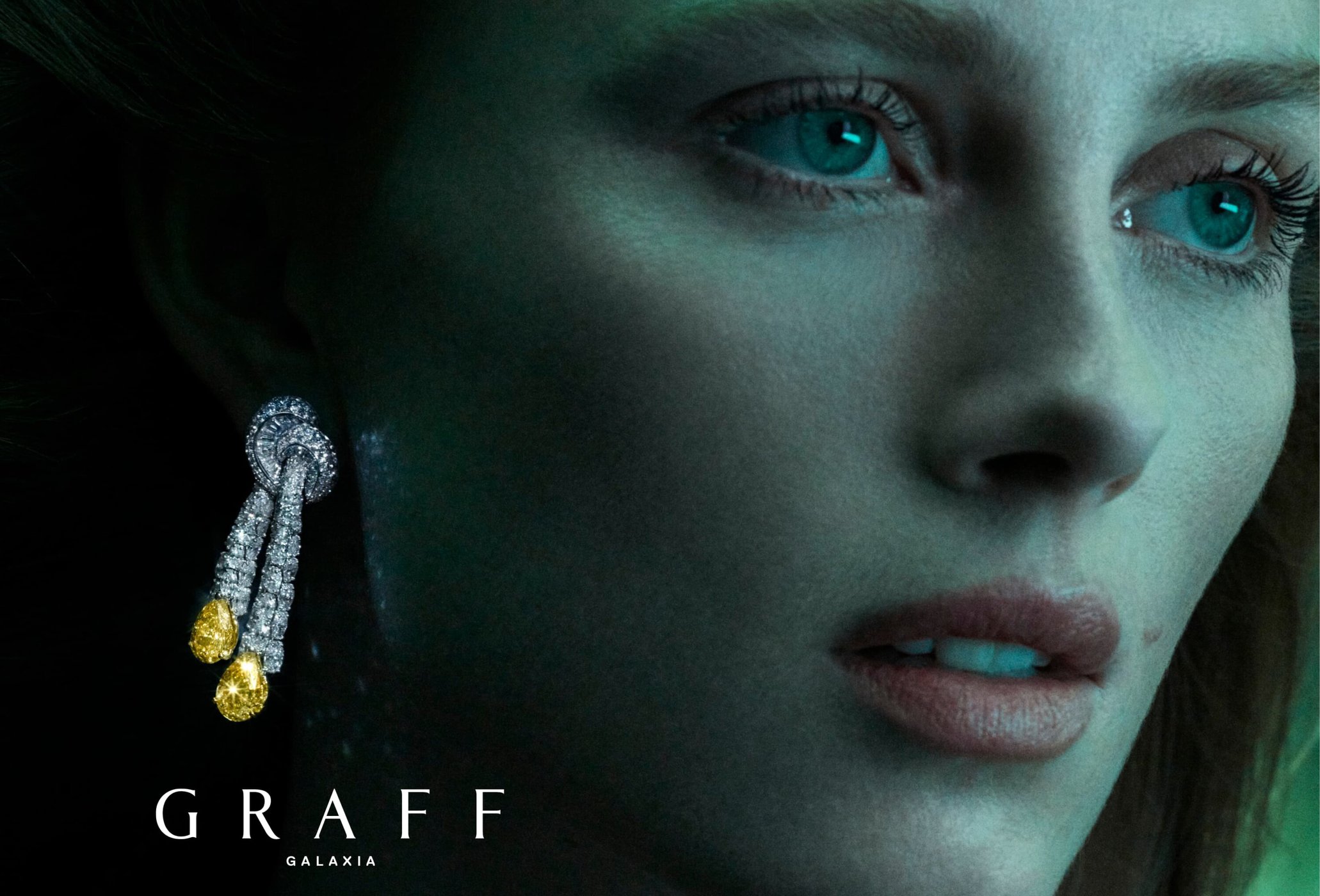 Close-up of an adult female model with blue eyes wearing Graff Galaxia diamond earrings against a dark green background.
