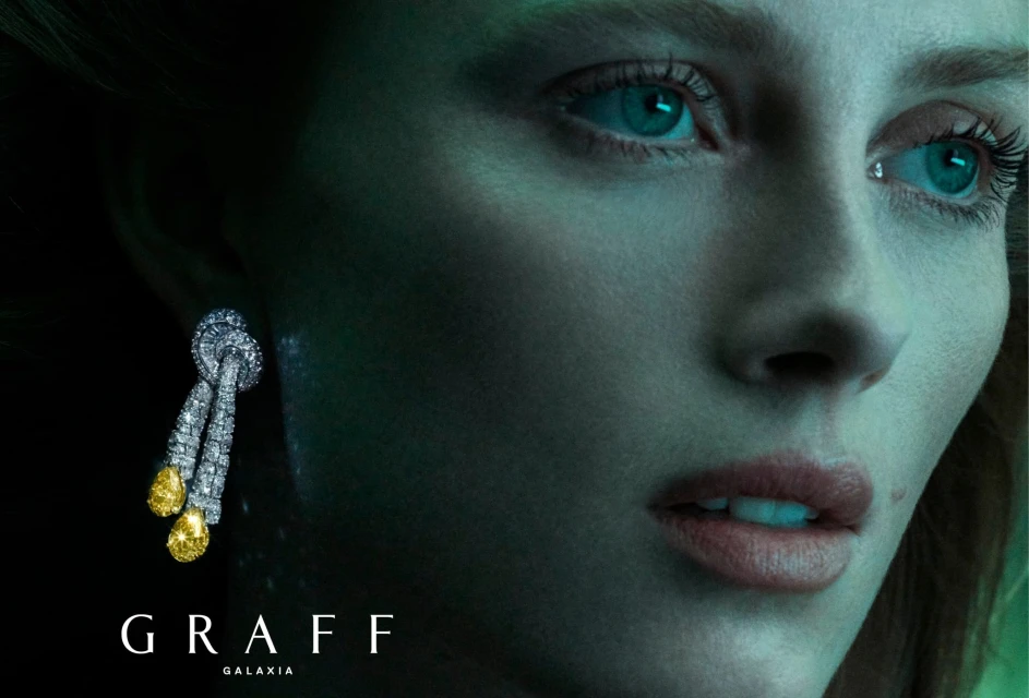 Close-up of an adult female model with blue eyes wearing Graff Galaxia diamond earrings against a dark green background.