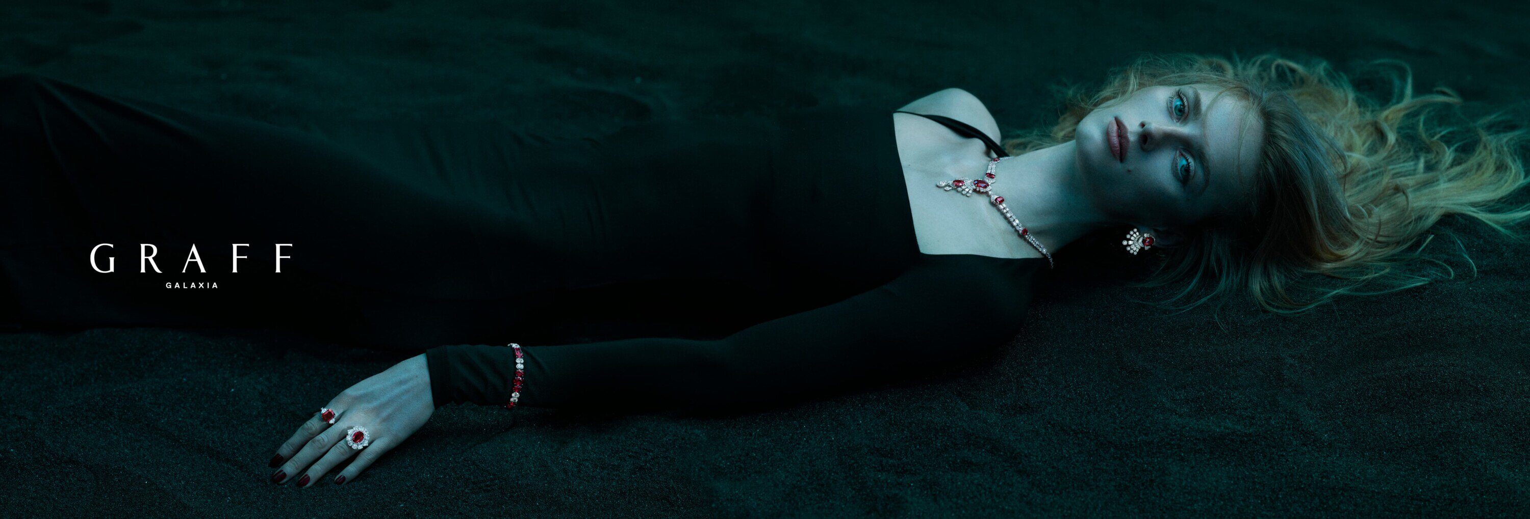 Model lying on dark sand wearing a black dress and Graff Galaxia jewelry including a necklace, bracelet, earrings, and ring.