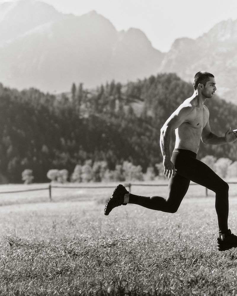 Monochrome image of an adult male runner in athletic wear sprinting across a grassy field with mountains in the background.