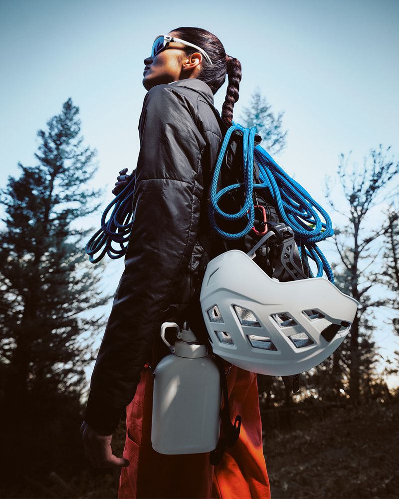Adult female hiker wearing a black puffer jacket, orange pants, and sunglasses carries climbing equipment in a forest setting.