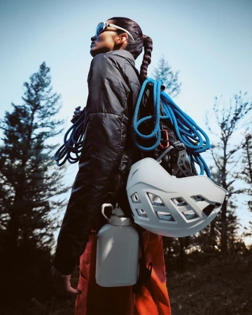 Adult female hiker wearing a black puffer jacket, orange pants, and sunglasses carries climbing equipment in a forest setting.