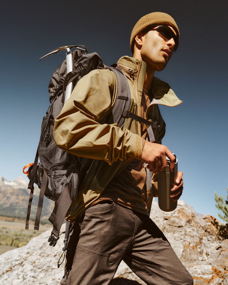 Male hiker opening a water bottle in the mountains, wearing a beanie and sunglasses, with a backpack and an ice axe attached.