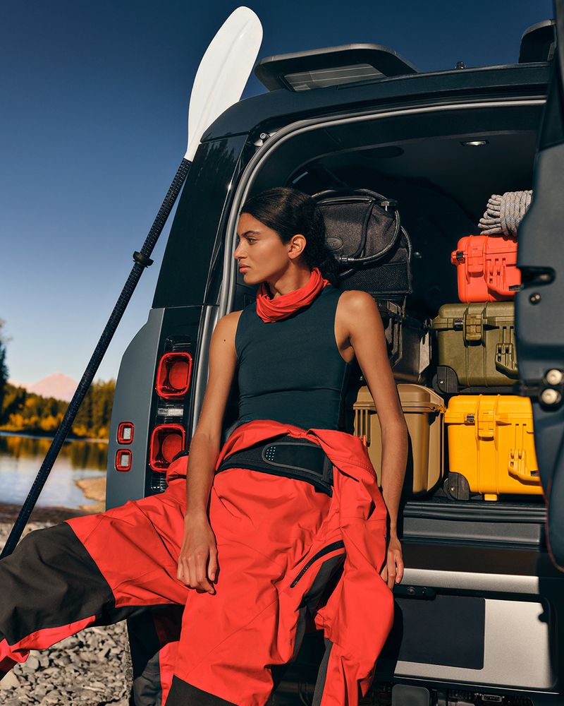 Woman in a wetsuit sits in the open back of a Land Rover Defender, ready for kayaking, with a paddle visible behind the vehicle.