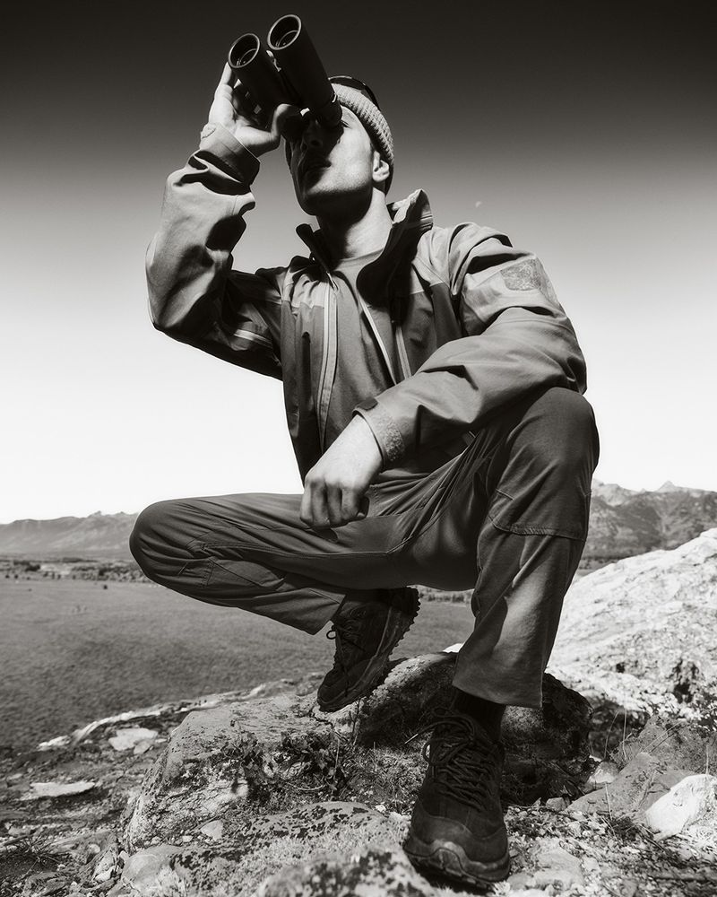 A man in outdoor gear squats on rocks, using binoculars in a monochrome outdoor shot with mountains in the background under a bright sky.