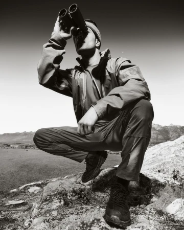 A man in outdoor gear squats on rocks, using binoculars in a monochrome outdoor shot with mountains in the background under a bright sky.