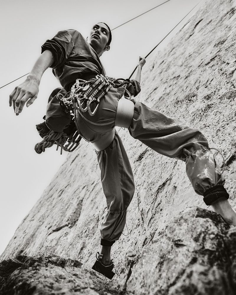 Monochrome, low-angle shot of a male rock climber ascending a textured rock face, secured by a rope and climbing gear.