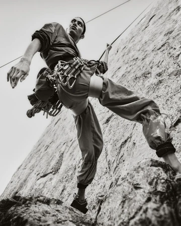 Monochrome, low-angle shot of a male rock climber ascending a textured rock face, secured by a rope and climbing gear.