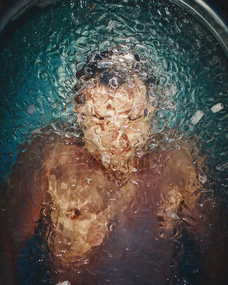 Underwater view of an adult with dark hair submerged in a tub of water and ice, captured with a high angle for a unique perspective.