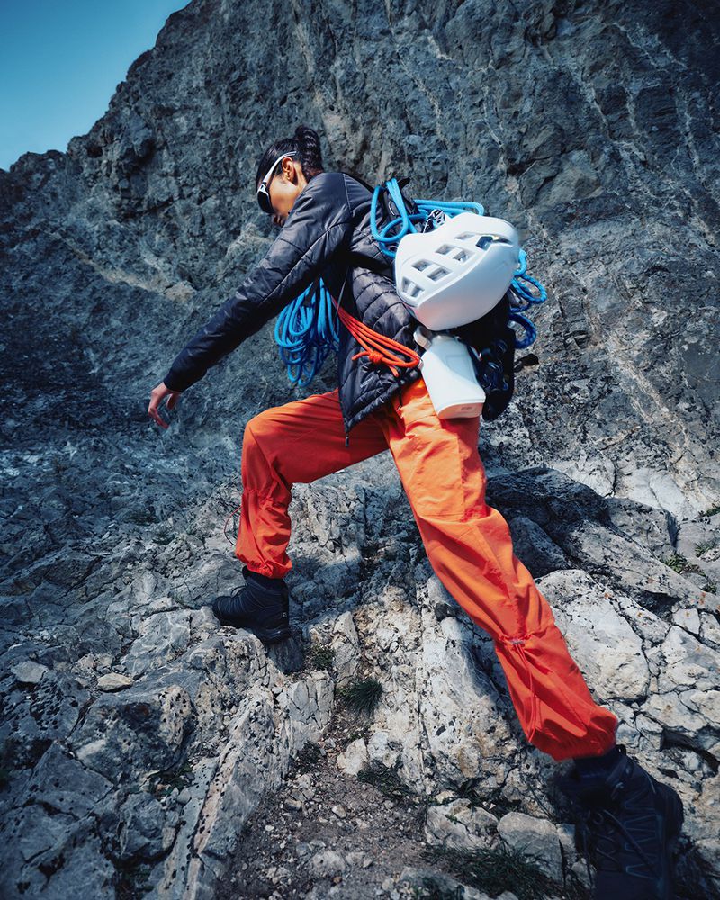 Female rock climber wearing a black jacket, orange pants, and climbing gear ascending a rocky mountain face on a sunny day.