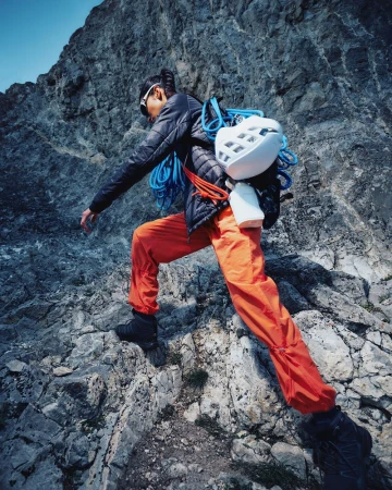 Female rock climber wearing a black jacket, orange pants, and climbing gear ascending a rocky mountain face on a sunny day.