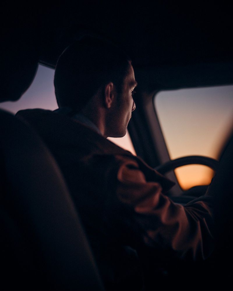 Adult male driving a car at dusk, seen from the backseat. Silhouette, neutral expression, warm sunset colors visible through windshield.