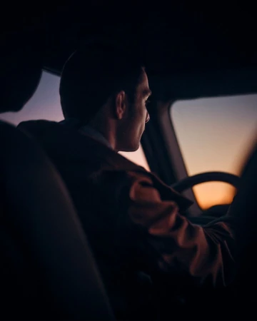 Adult male driving a car at dusk, seen from the backseat. Silhouette, neutral expression, warm sunset colors visible through windshield.