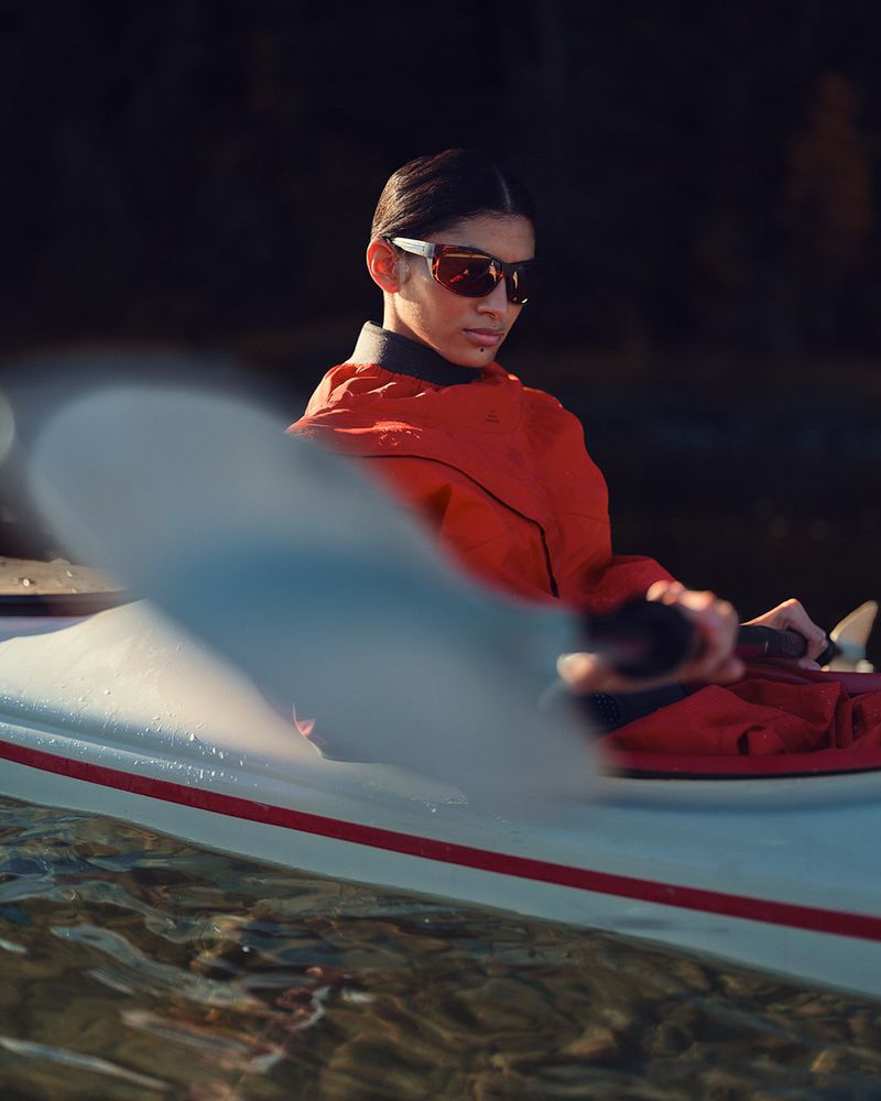 A woman in sunglasses and a red jacket paddles a white kayak on water in a dark outdoor setting, showcasing sport and outdoor activity.
