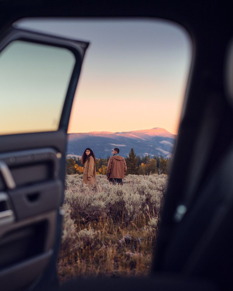 A couple stands in a grassy field with mountains in the background, viewed through an open car door at sunset.