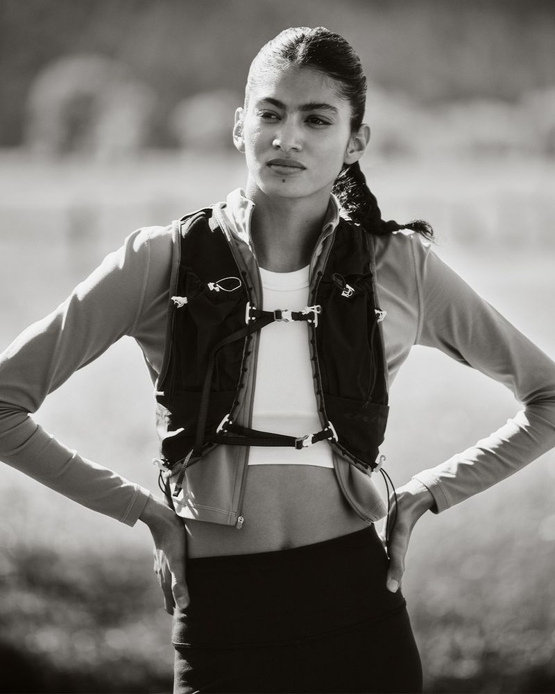 Monochrome shot of a woman in activewear and a vest, standing outdoors with her hands on her hips, looking off into the distance.