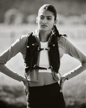Monochrome shot of a woman in activewear and a vest, standing outdoors with her hands on her hips, looking off into the distance.