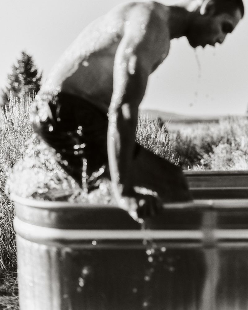 Black and white photo of an adult male sitting in a metal water trough outside, water dripping from his face.