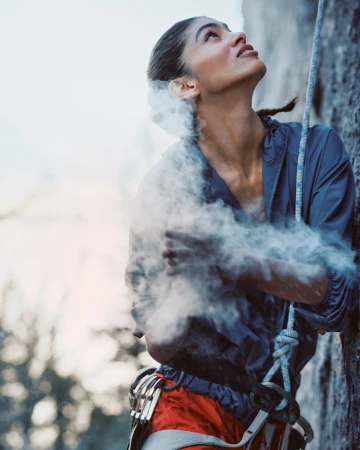 Female rock climber pauses on a rock face dusted with chalk, looking up with determination, wearing outdoor clothing and climbing harness.