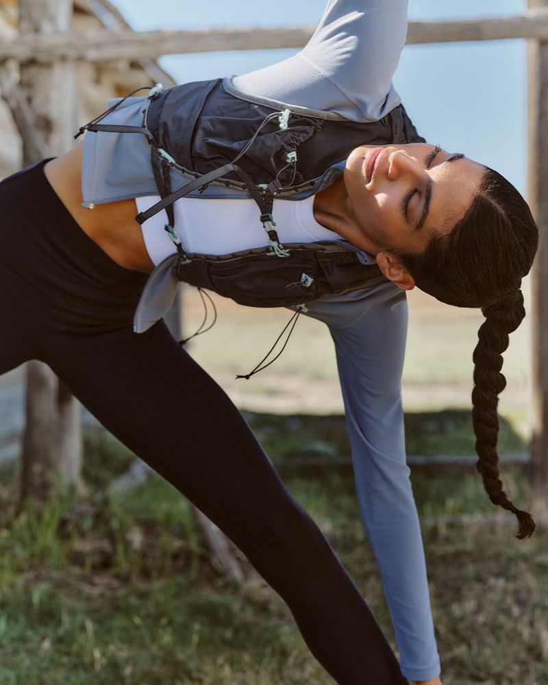 Woman in black leggings and athletic top poses in yoga stance outdoors with light filtering through trees. Hair is braided.