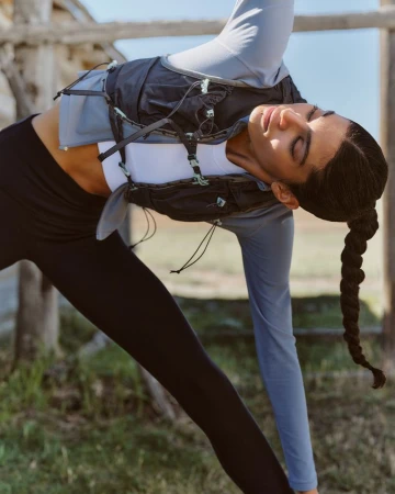 Woman in black leggings and athletic top poses in yoga stance outdoors with light filtering through trees. Hair is braided.