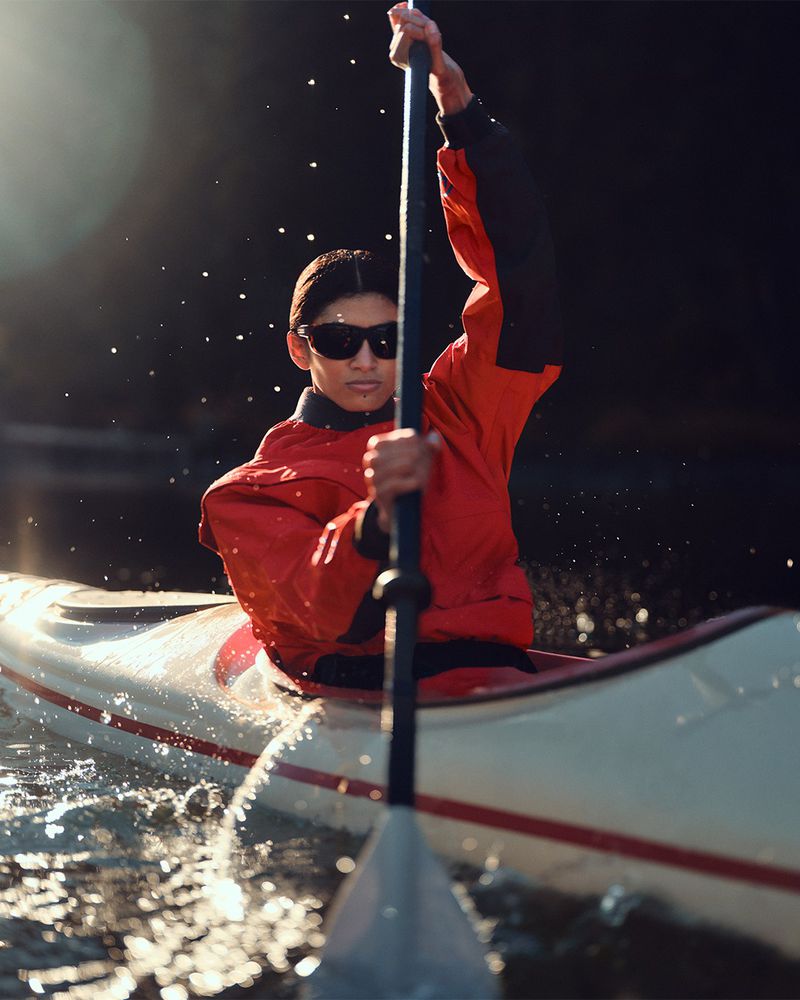 An adult female in a red jacket paddles a kayak on sunlit water with a raised paddle and sunglasses on a sunny day for sport.