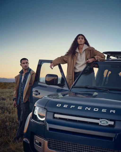 A navy Land Rover Defender is parked outdoors with a man and woman casually posed by it at dusk in a lifestyle fashion shoot.