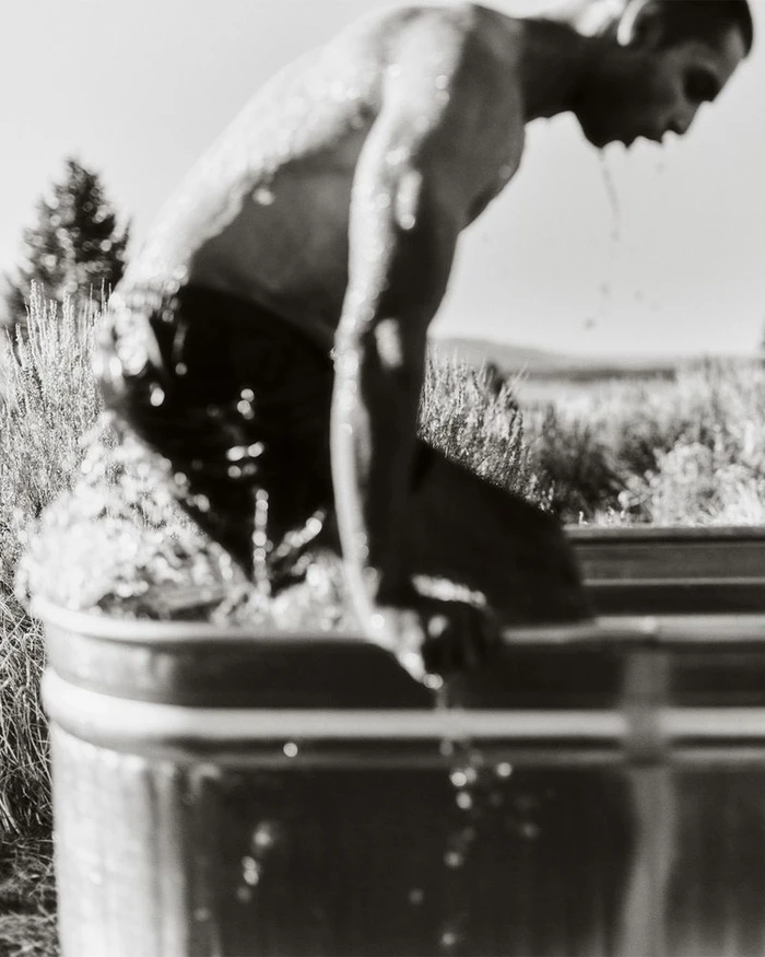 Monochrome action shot of a man climbing into a galvanized metal stock tank outdoors amidst tall grasses.