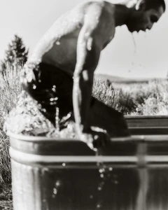 Monochrome action shot of a man climbing into a galvanized metal stock tank outdoors amidst tall grasses.