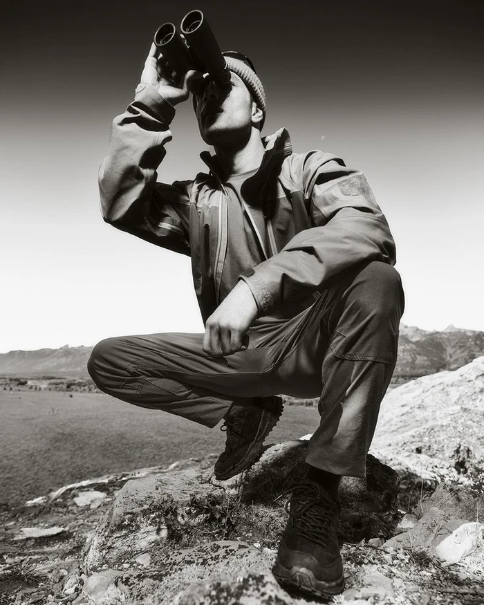 Man crouching on rocky terrain holding binoculars to his eyes against an open sky for Land Rover Defender Wyoming 2025 campaign.