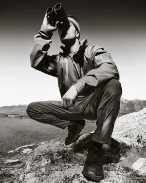 Man crouching on rocky terrain holding binoculars to his eyes against an open sky for Land Rover Defender Wyoming 2025 campaign.