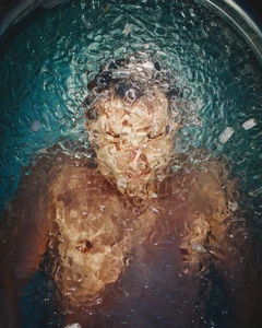 Model submerged underwater surrounded by floating ice cubes and rippling blue water in an overhead portrait shot.