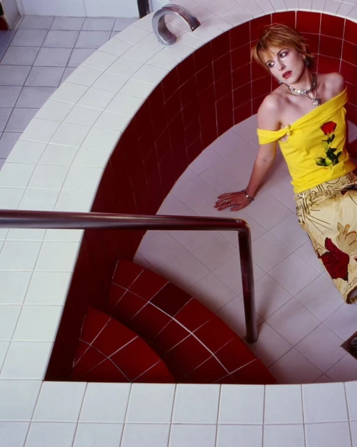 Adult female model in yellow top with floral pants poses in a bathroom setting with red and white tiles, metal railings and fixtures.