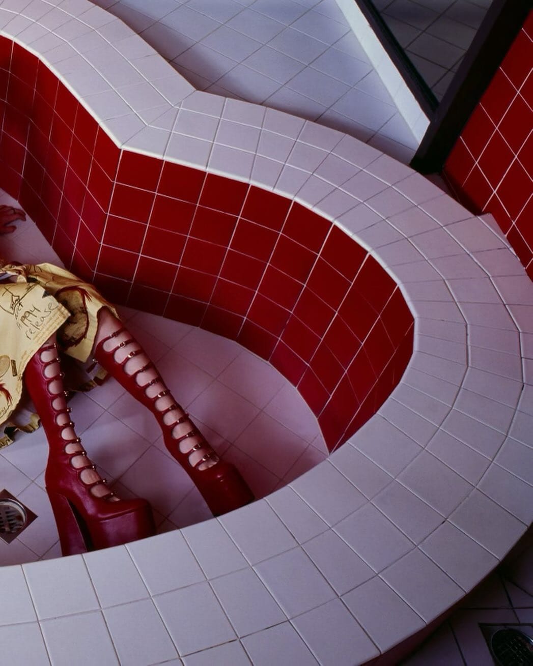 Fashion photo of a person wearing red platform boots in a geometric, tiled space with red and white square tiles, high-angle view.