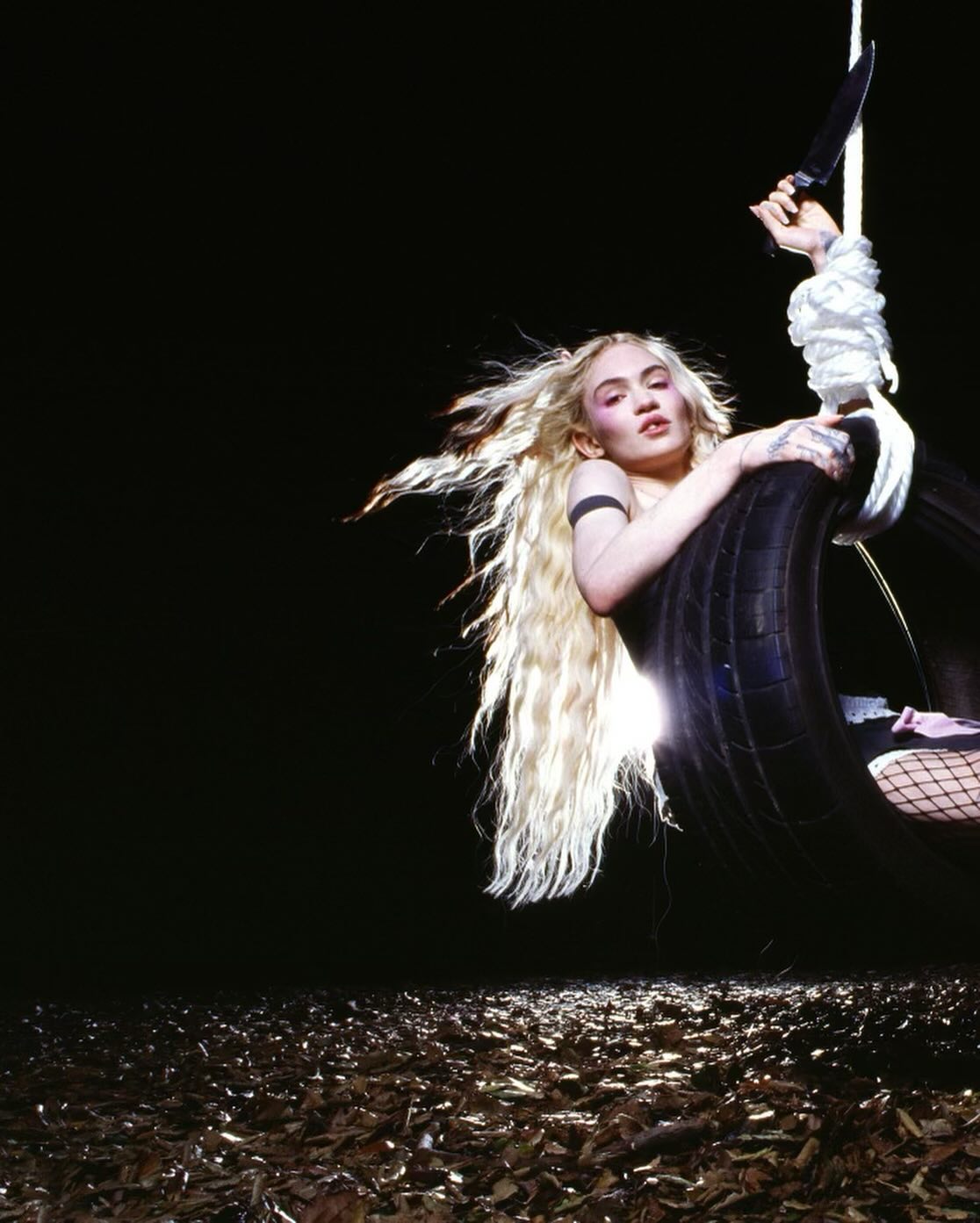 Portrait of a blonde adult woman in a tire swing, holding a knife, in an outdoor setting with leaves on the ground and a black background.