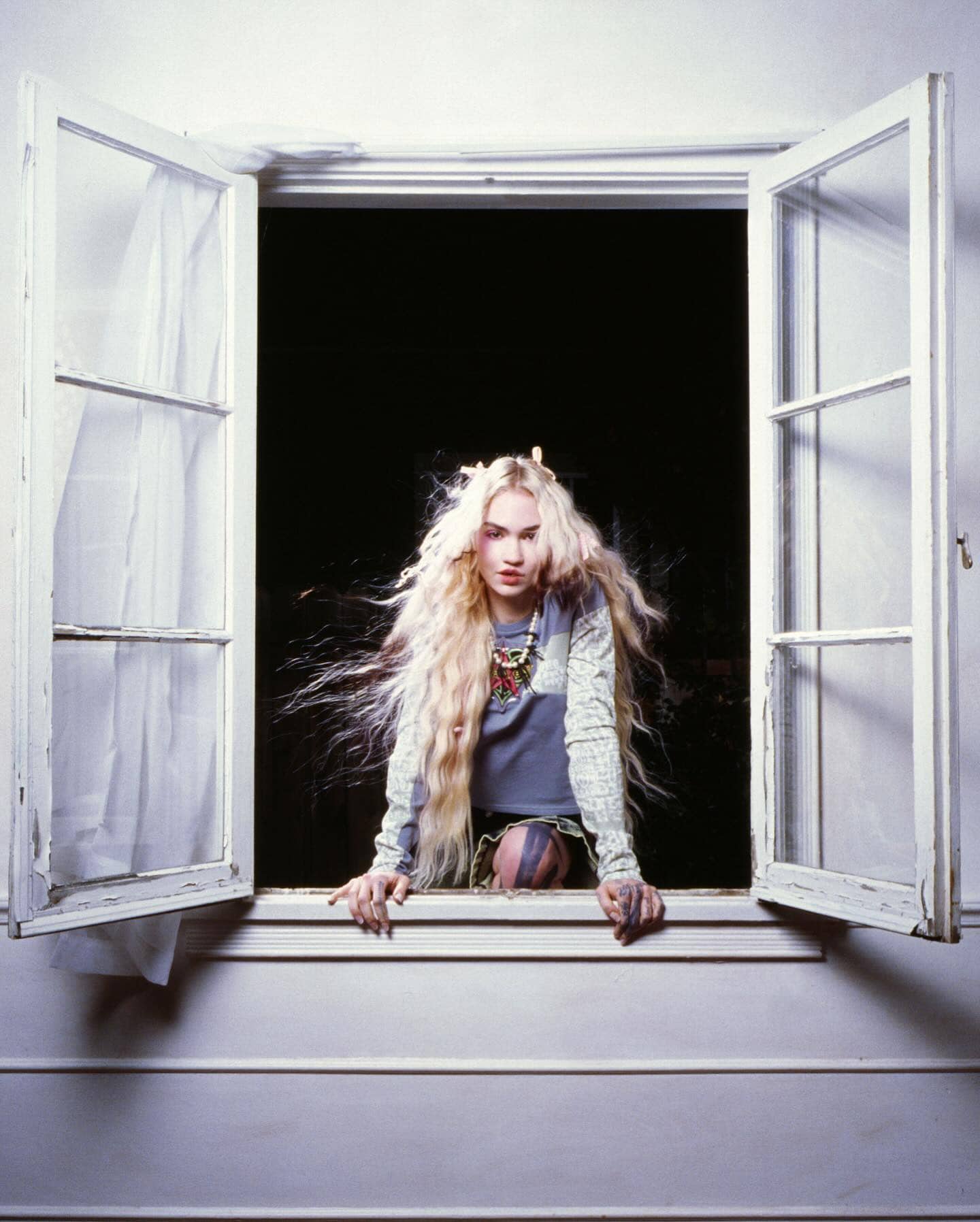 A fair-skinned young adult female with long blonde hair is shown at a white-framed open window with a black backdrop, looking at the viewer.