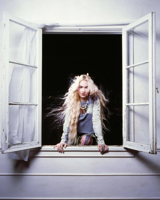 A fair-skinned young adult female with long blonde hair is shown at a white-framed open window with a black backdrop, looking at the viewer.
