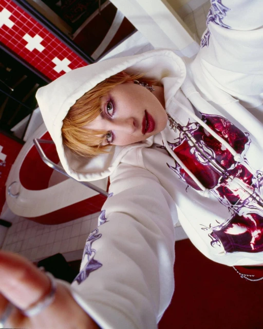 Young woman in white graphic hoodie poses in a low-angle shot with a red and white patterned background, showcasing urban fashion.