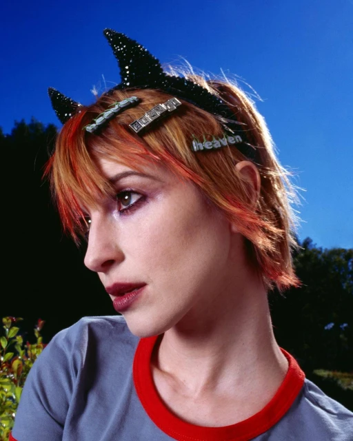 Portrait of a young adult female with dyed orange hair, devil horn headband, and hair clips against a blue sky and greenery background.