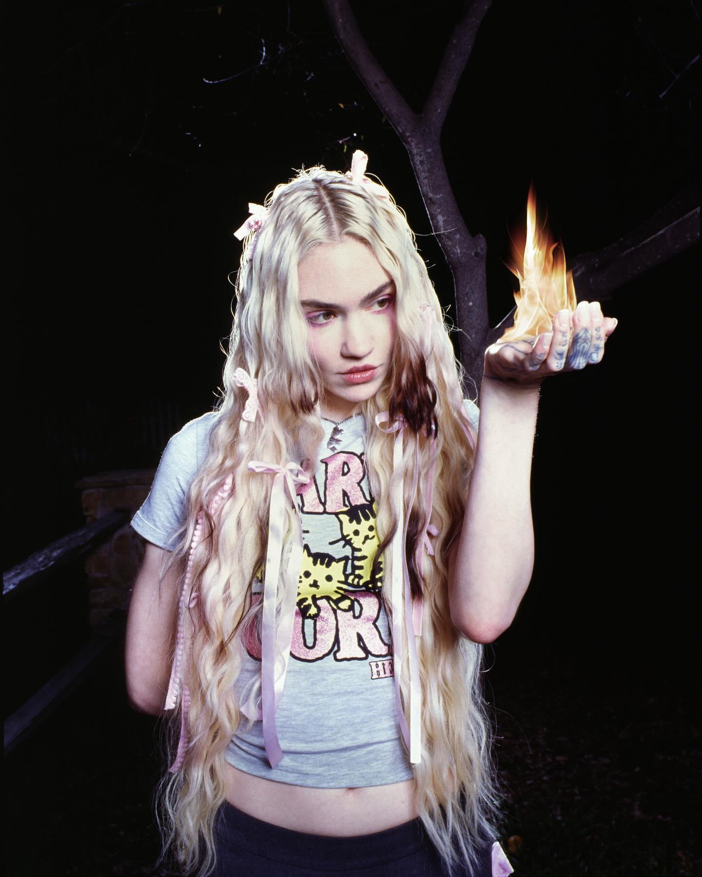 Young woman holding flames in her hand, wearing a graphic t-shirt and hair ribbons. Dark background with tree branches.