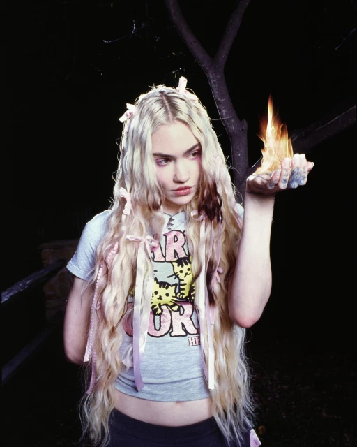 Young woman holding flames in her hand, wearing a graphic t-shirt and hair ribbons. Dark background with tree branches.