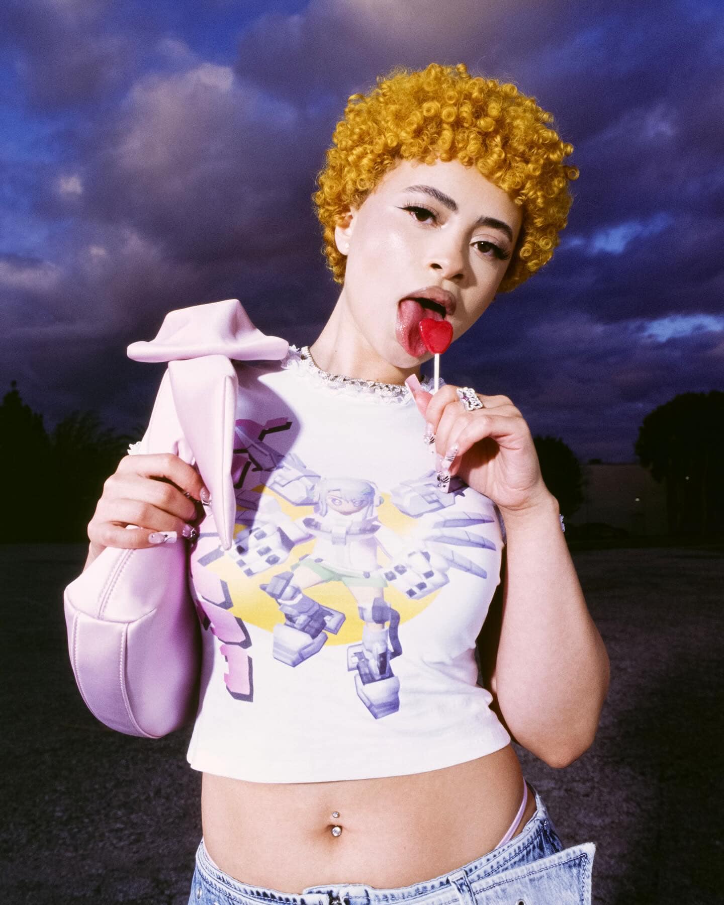 Young woman with curly blonde hair poses outdoors with a heart lollipop. She wears a graphic tee, low-rise jeans, and a pink purse.