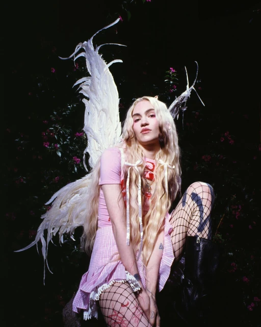Young woman in fairy costume with feathered wings poses in an outdoor garden with dark lighting and pink flowers in the background.